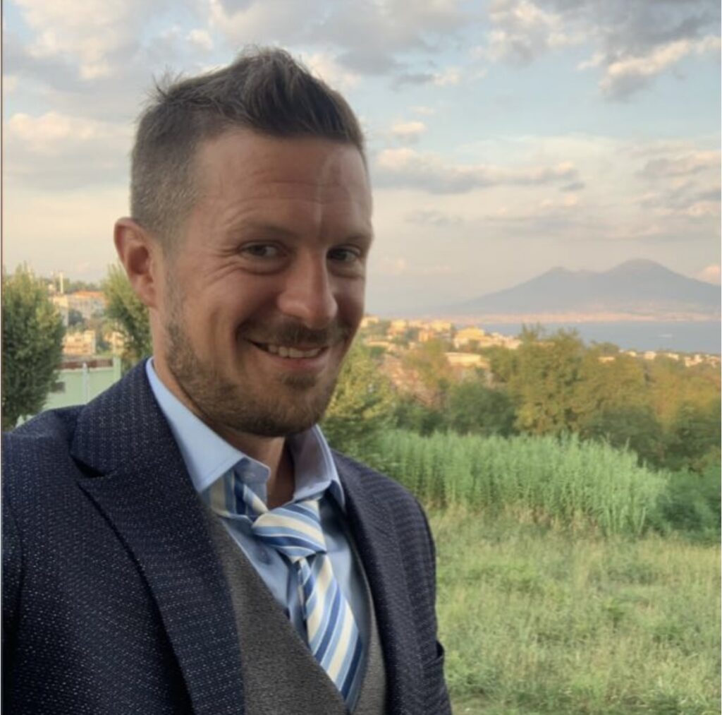 Emilio sorridente guarda verso l'obiettivo della telecamera e alle spalle ha un paesaggio verde e una montagna all'orizzonte.